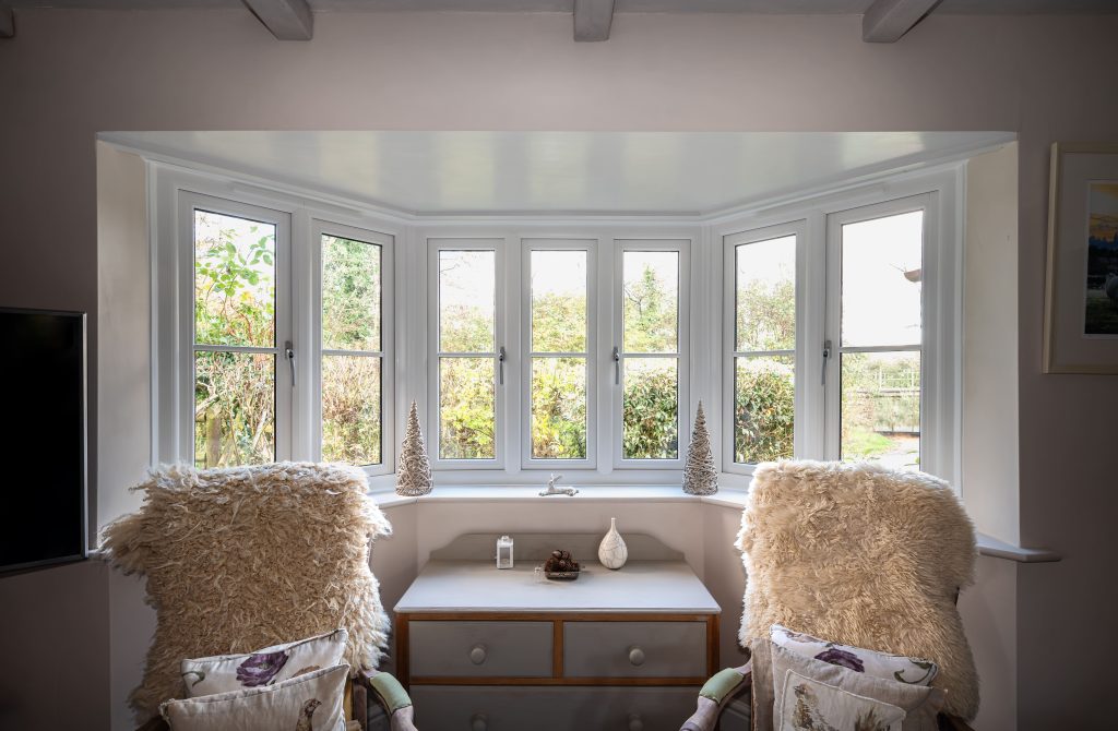 A bright bay window with white frames, overlooking a garden. Two cozy armchairs with fluffy cushions face the window, with a small chest of drawers between them, decorated with ornaments.